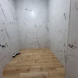 Wykończona łazienka z białymi płytkami imitującymi marmur na ścianach i drewnopodobną podłogą. Widoczne rury instalacyjne, gotowe do podłączenia armatury.