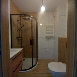 Usługi Remontowo Budowlane Rob-bud - Nowoczesna łazienka z kabiną prysznicową typu walk-in, umywalką z drewnianą szafką i podwieszaną toaletą. Biało-drewniane wykończenie i minimalistyczne oświetlenie.