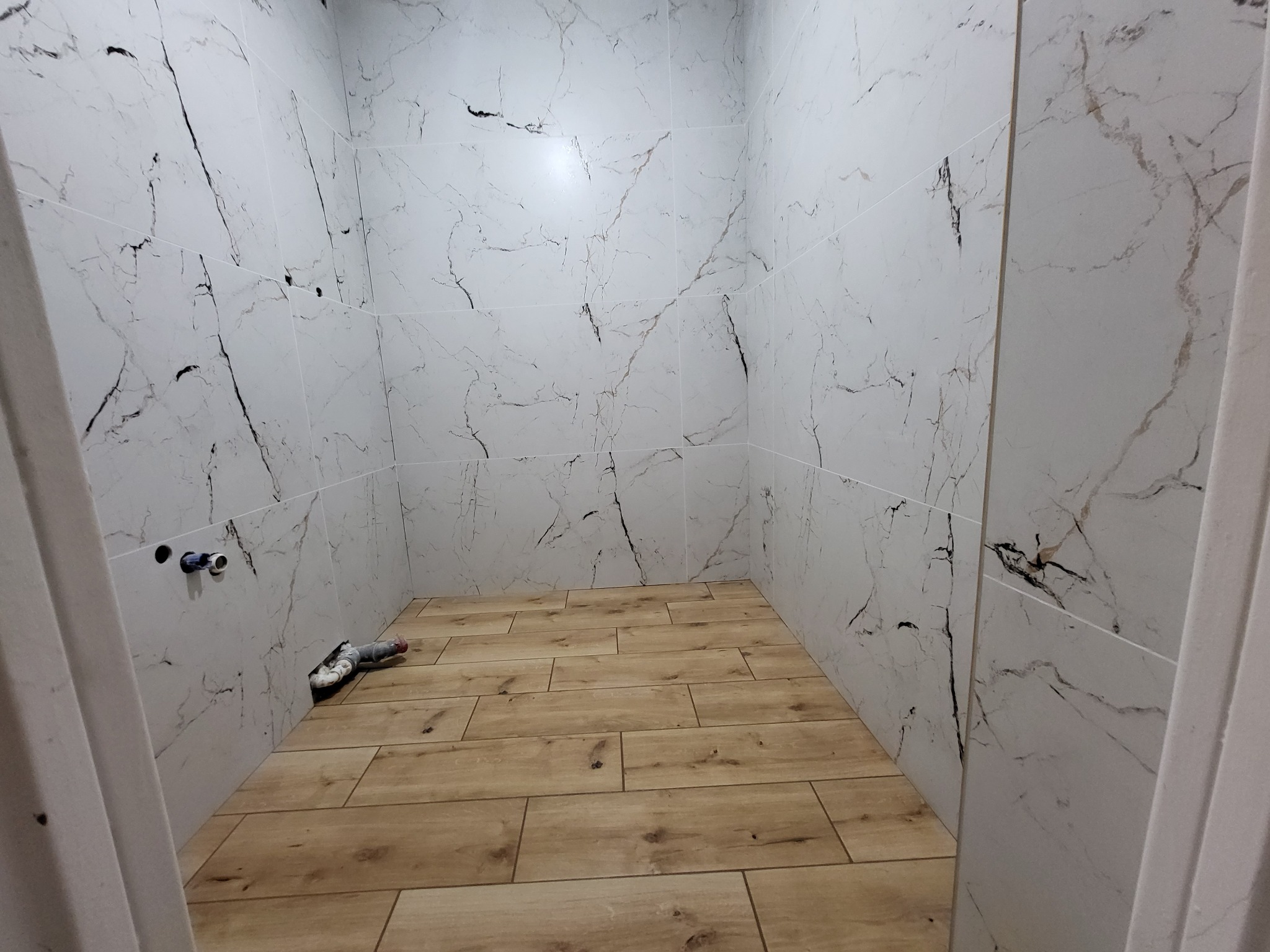 Wykończona łazienka z białymi płytkami imitującymi marmur na ścianach i drewnopodobną podłogą. Widoczne rury instalacyjne, gotowe do podłączenia armatury.