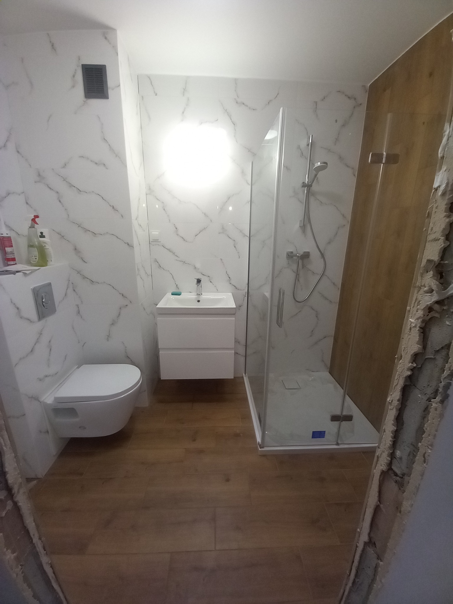 Wykończona łazienka z kabiną prysznicową, toaletą i umywalką. Ściany wyłożone białymi płytkami imitującymi marmur, podłoga drewnopodobna. Widoczne niedokończone wykończenie przy drzwiach.