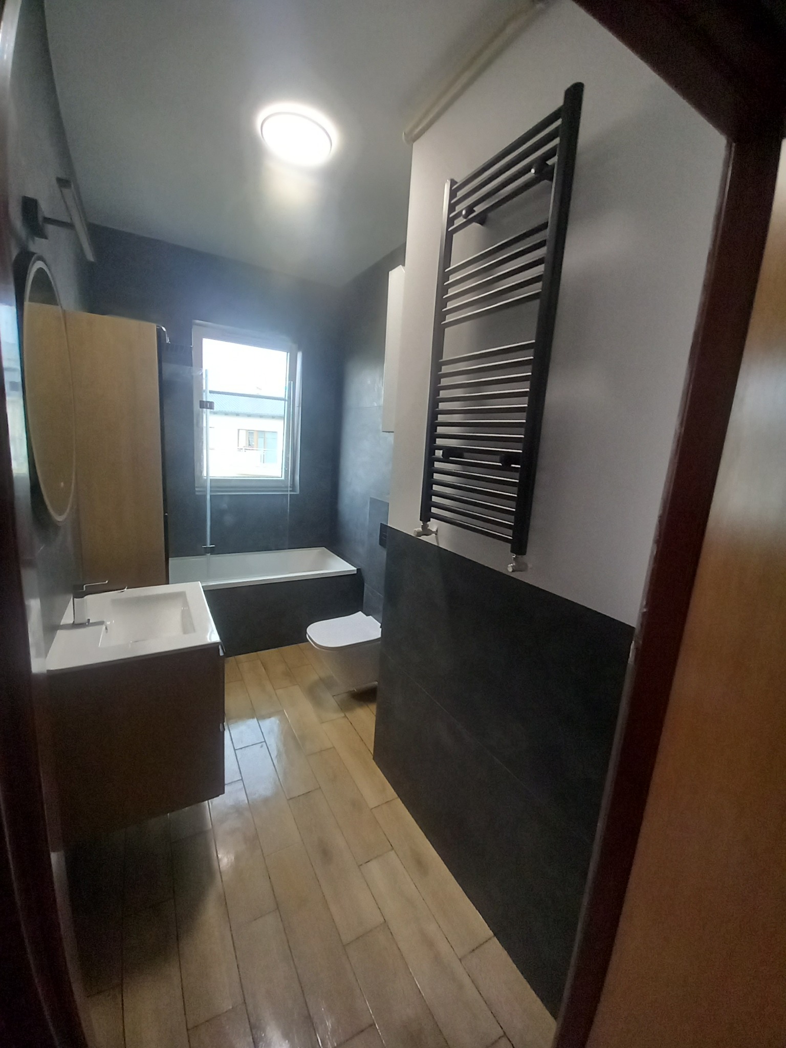 Nowoczesna łazienka z drewnopodobną podłogą, grafitowymi ścianami i grzejnikiem drabinkowym. Widok na umywalkę, wannę i toaletę. Minimalistyczny design.