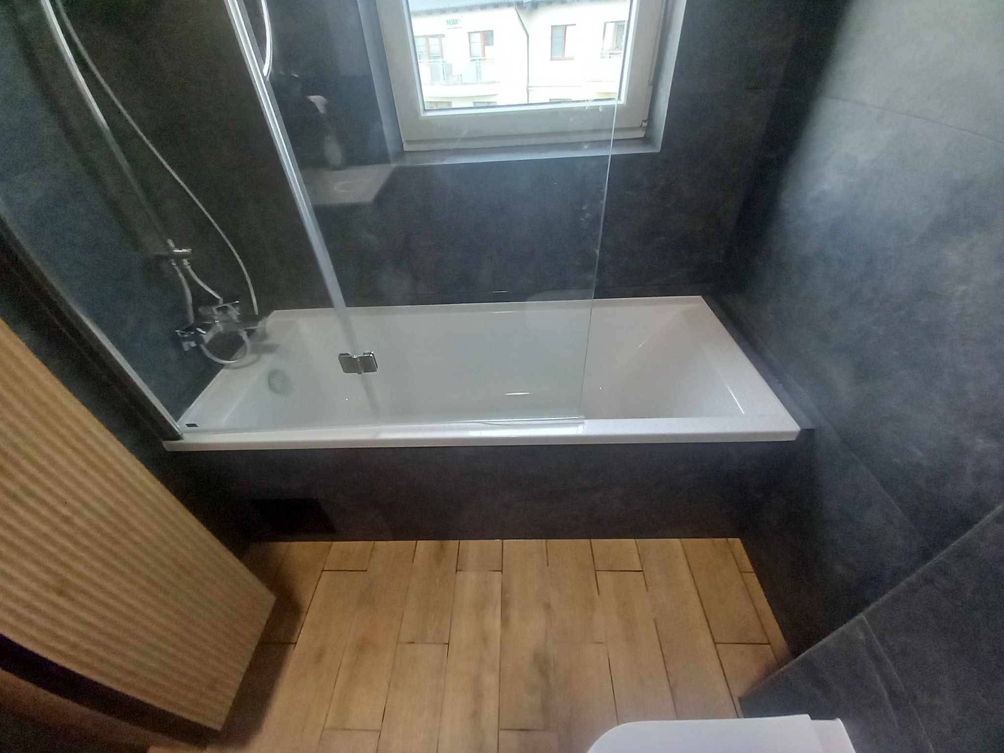 Nowoczesna łazienka z wanną pod oknem, wykończona w szarościach z drewnianą podłogą. Kabina prysznicowa ze szklaną ścianką.