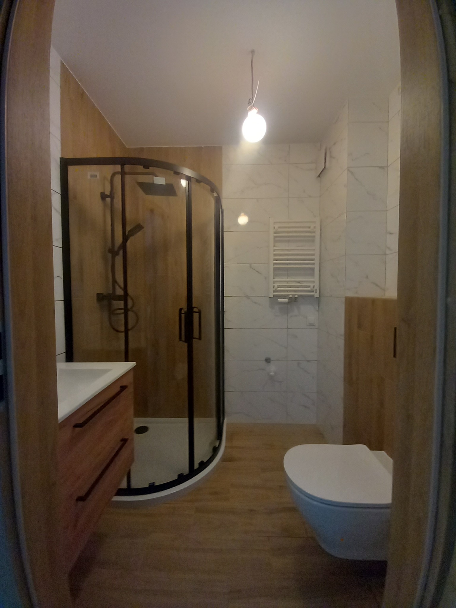 Nowoczesna łazienka z kabiną prysznicową typu walk-in, umywalką z drewnianą szafką i podwieszaną toaletą. Biało-drewniane wykończenie i minimalistyczne oświetlenie.