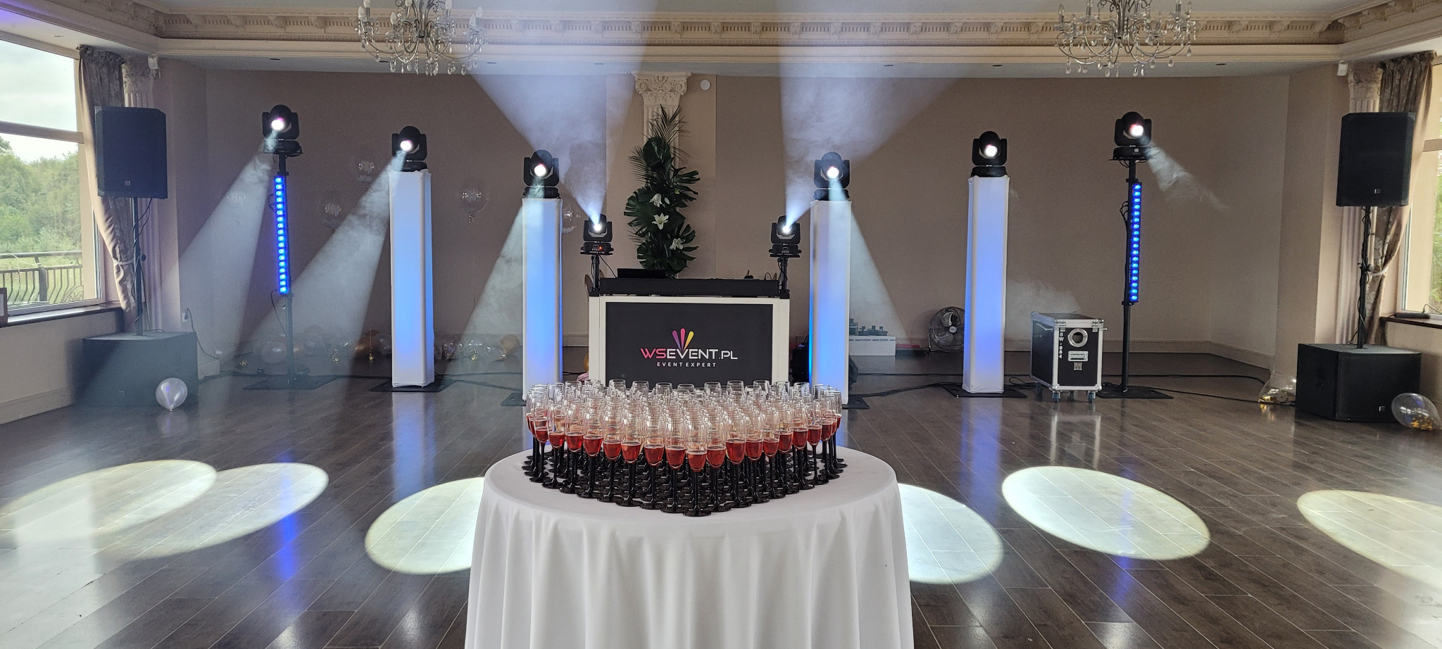 Ustawiony sprzęt nagłośnieniowy i oświetleniowy z logo firmy WS Event.pl, stół z piramidą kieliszków wypełnionych czerwonym płynem, dekoracja sali z balonami.
