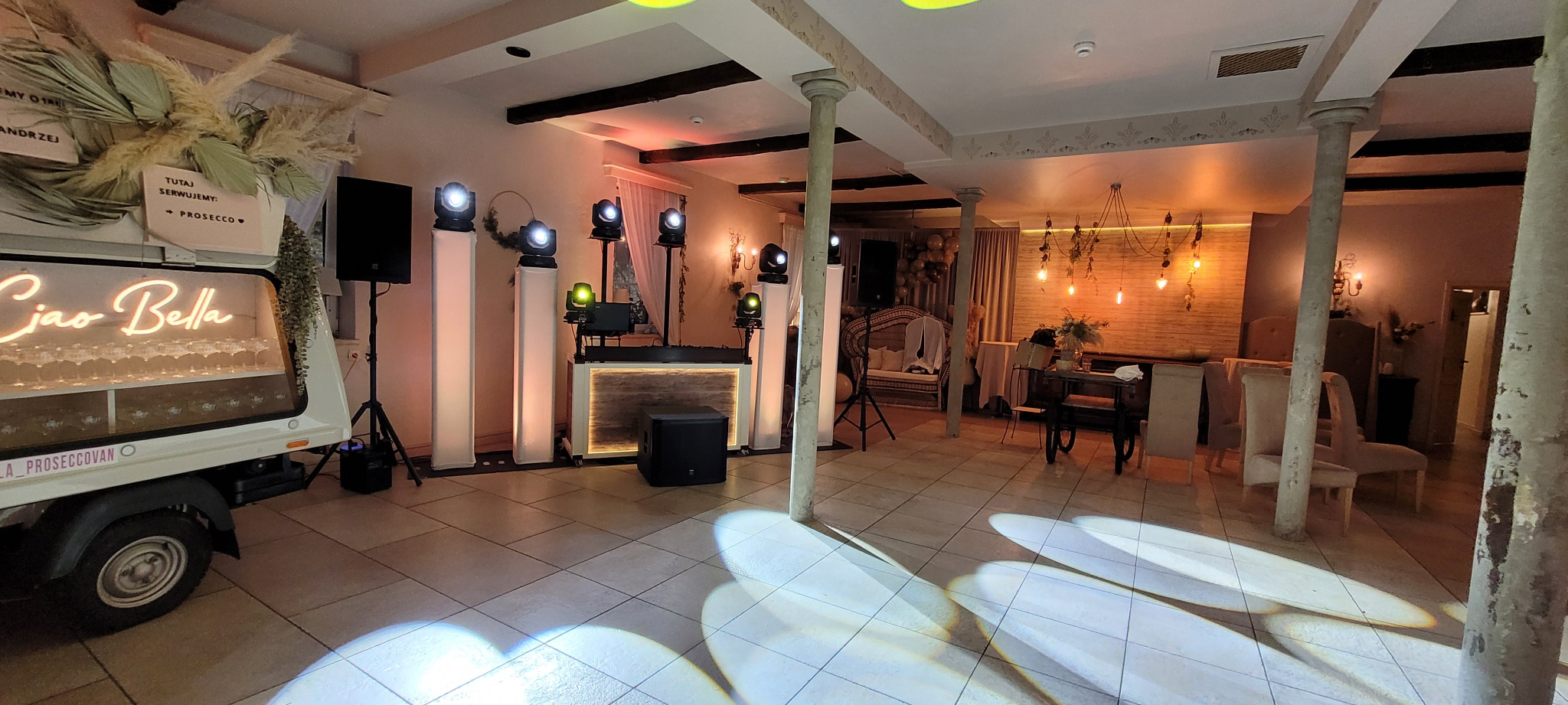 Sala bankietowa z oświetleniem eventowym, barem prosecco na kółkach, kolumnami i dekoracjami roślinnymi.