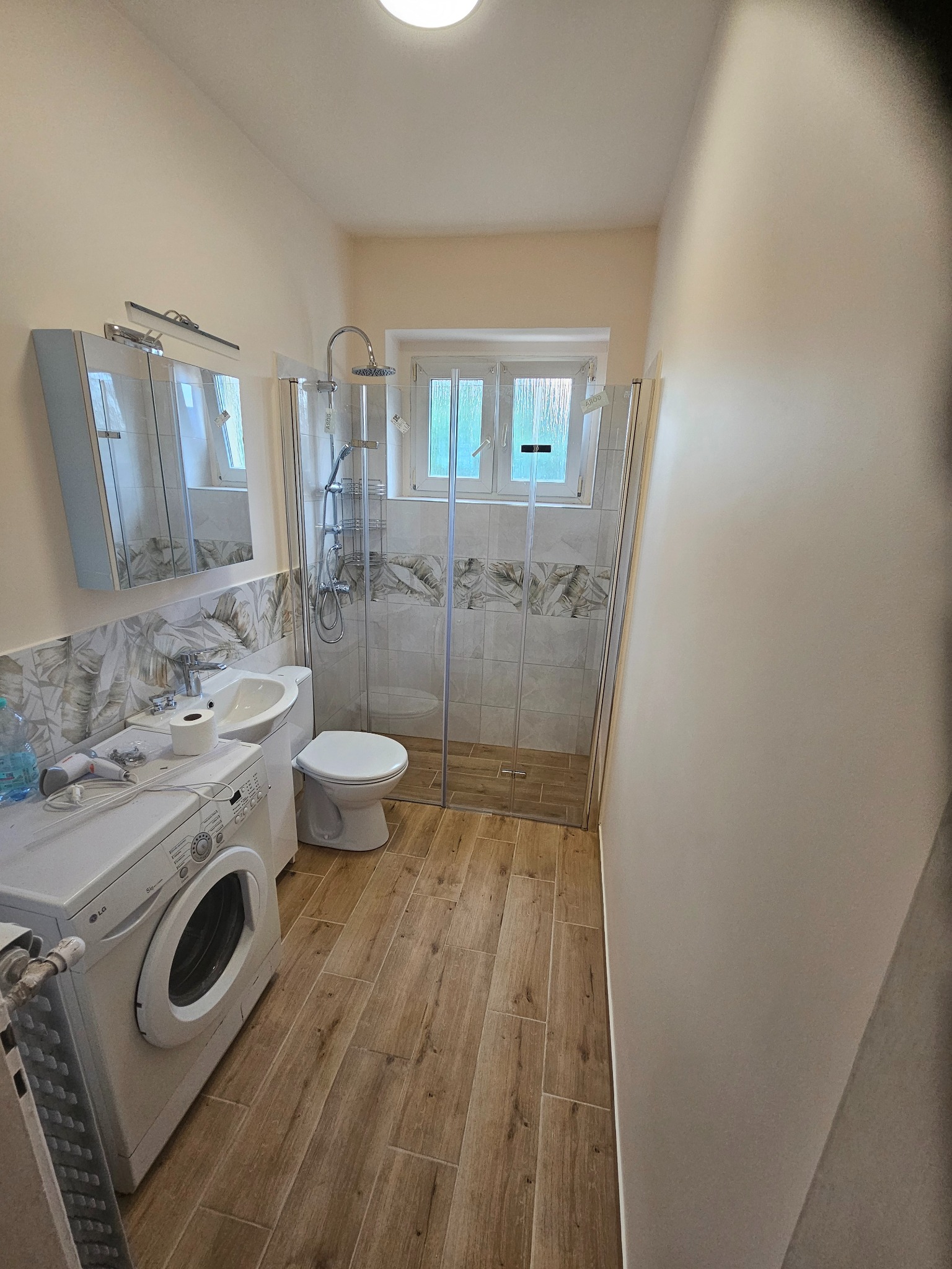 Wąska łazienka po remoncie z kabiną prysznicową, umywalką z szafką lustrzaną, toaletą i pralką LG, wykończona płytkami imitującymi drewno i dekoracyjnymi płytkami z motywem liści.