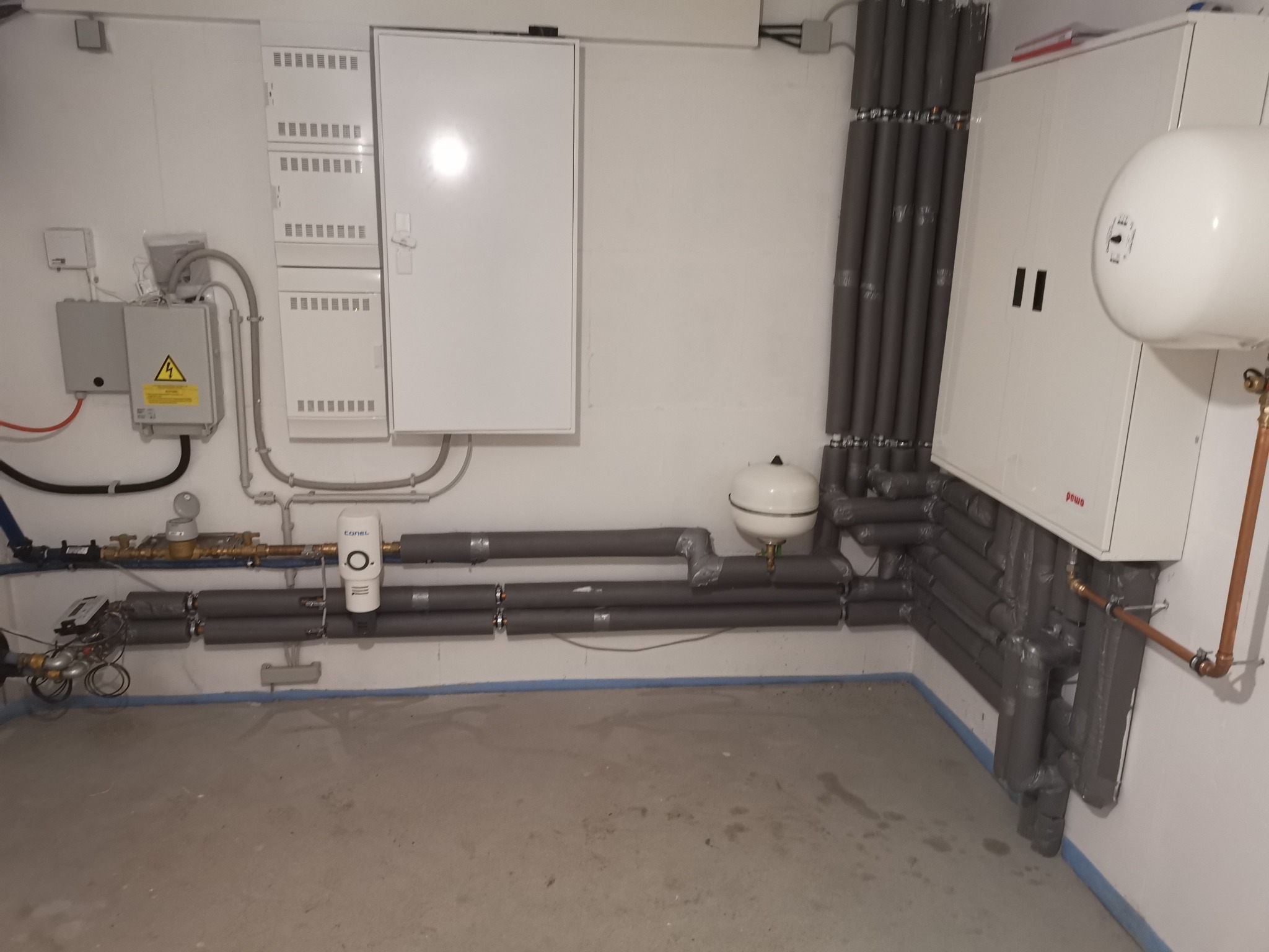 Piwnica z instalacją grzewczą: rury w izolacji, biały zbiornik wyrównawczy, szafka sterownicza i rozdzielnica elektryczna na ścianie.