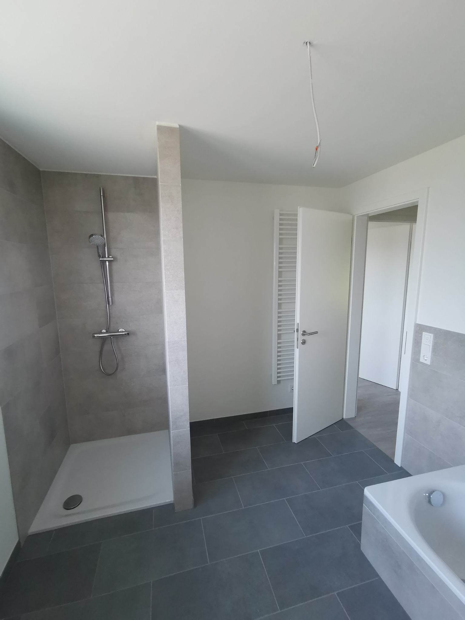 Nowoczesna łazienka w szarościach, z kabiną prysznicową typu walk-in, grzejnikiem drabinkowym i częściowo zamontowaną wanną, widoczne przewody elektryczne wystające z sufitu wskazują na trwające...