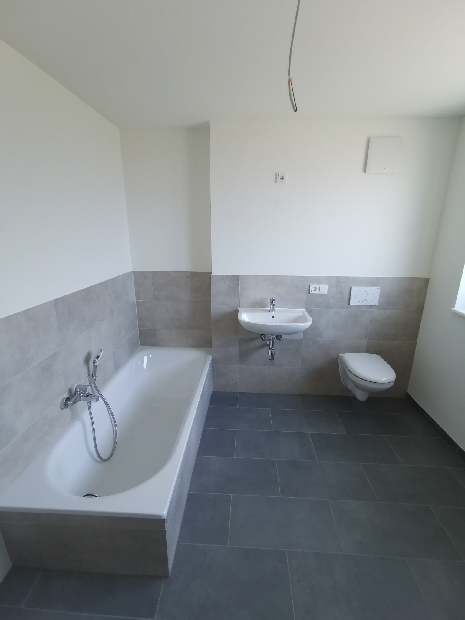 Nowoczesna łazienka po remoncie z szarymi płytkami, białą wanną, umywalką i toaletą, widoczne przewody elektryczne na suficie.