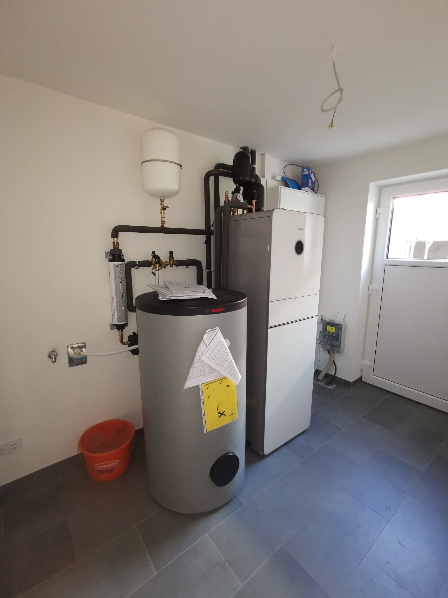 Nowoczesna kotłownia z szarym zasobnikiem ciepłej wody, białym piecem kondensacyjnym Bosch i pomarańczowym wiadrem na szarej podłodze z płytek.