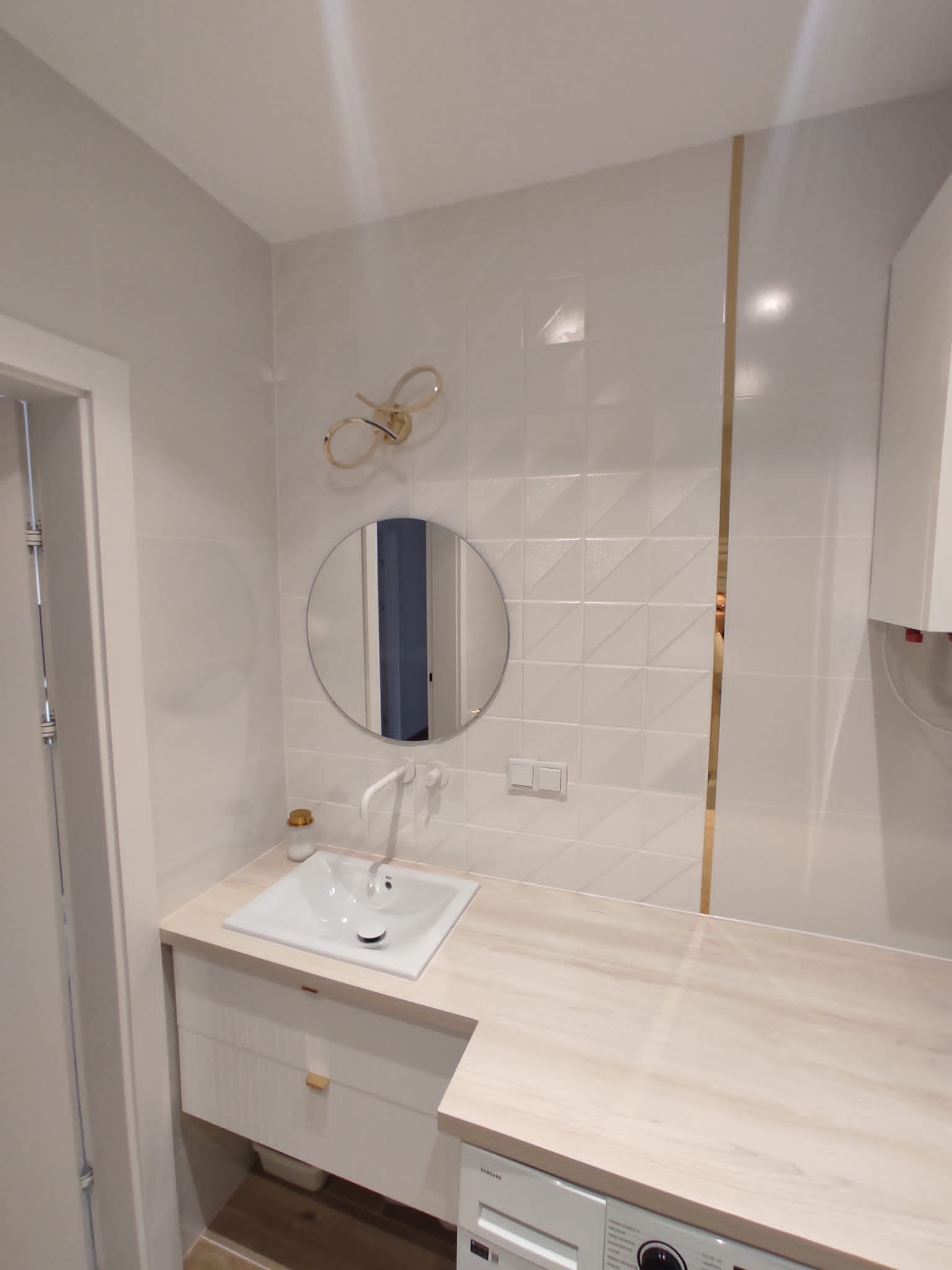 Nowoczesna łazienka z okrągłym lustrem, białą umywalką i blatem imitującym drewno. Na ścianie białe, geometryczne płytki z złotym akcentem. Widoczny fragment pralki.