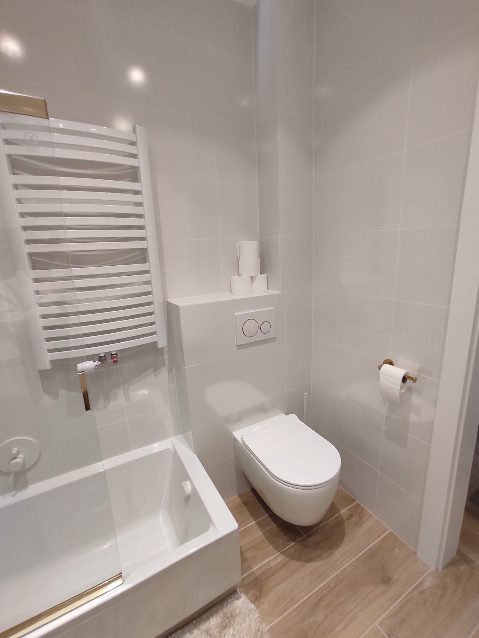 Nowoczesna łazienka z białymi płytkami, podwieszaną toaletą, grzejnikiem drabinkowym i drewnopodobną podłogą. Minimalistyczny design z akcentami złota.