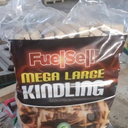 Mskills Spółka z o.o. - Worek z przezroczystego tworzywa wypełniony drobnymi kawałkami drewna opałowego, etykieta FuelSell Mega Large Kindling, drewniane palety w tle.
