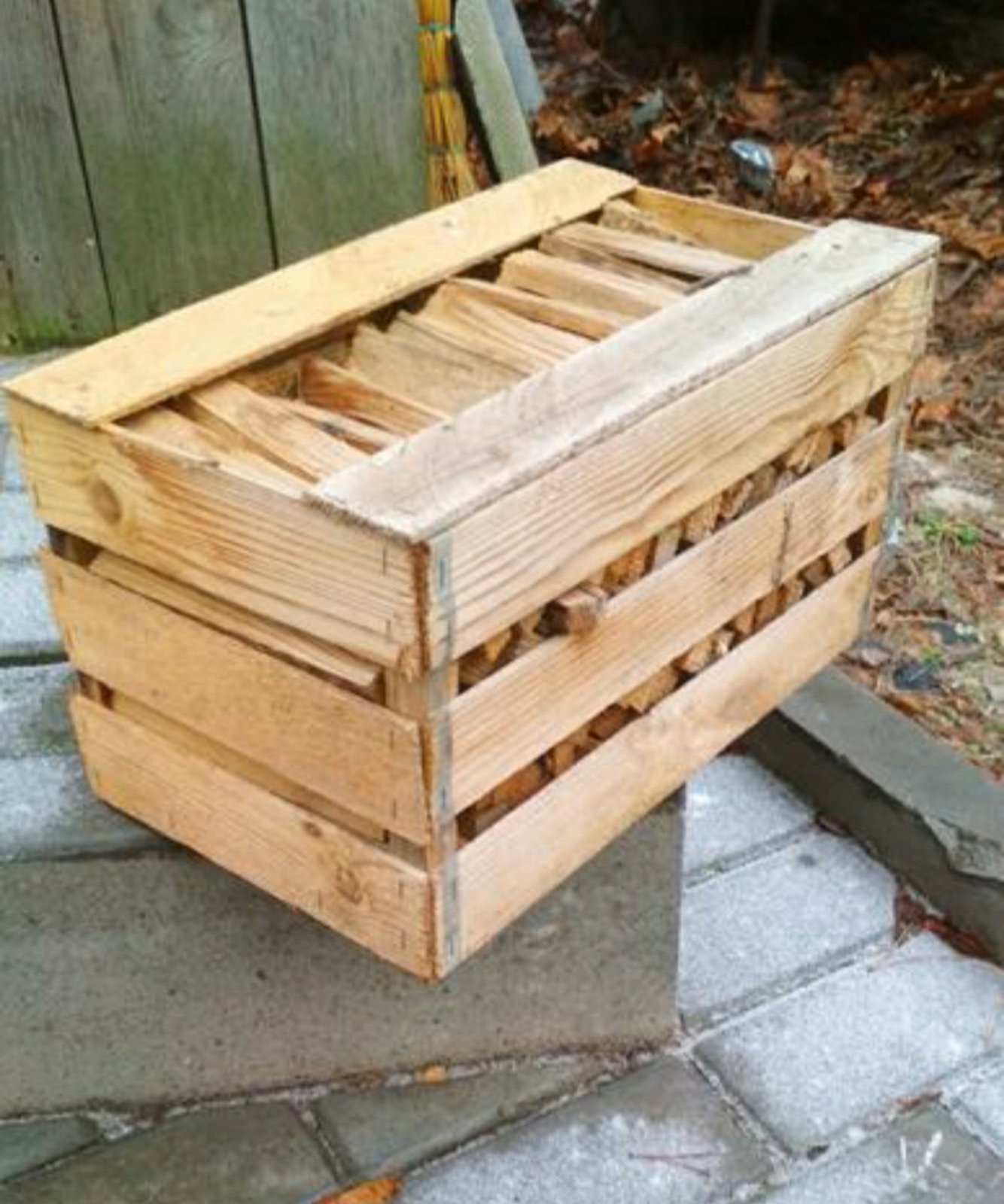 Drewniana skrzynia wypełniona pociętym drewnem kominkowym, ustawiona na szarych betonowych płytach chodnikowych, w tle widoczna drewniana ściana i liście.