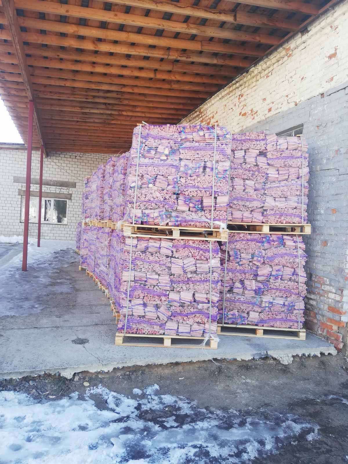 Stosy palet z drewnem kominkowym, zabezpieczonym fioletową folią, składowane pod zadaszeniem przy ceglanym budynku.