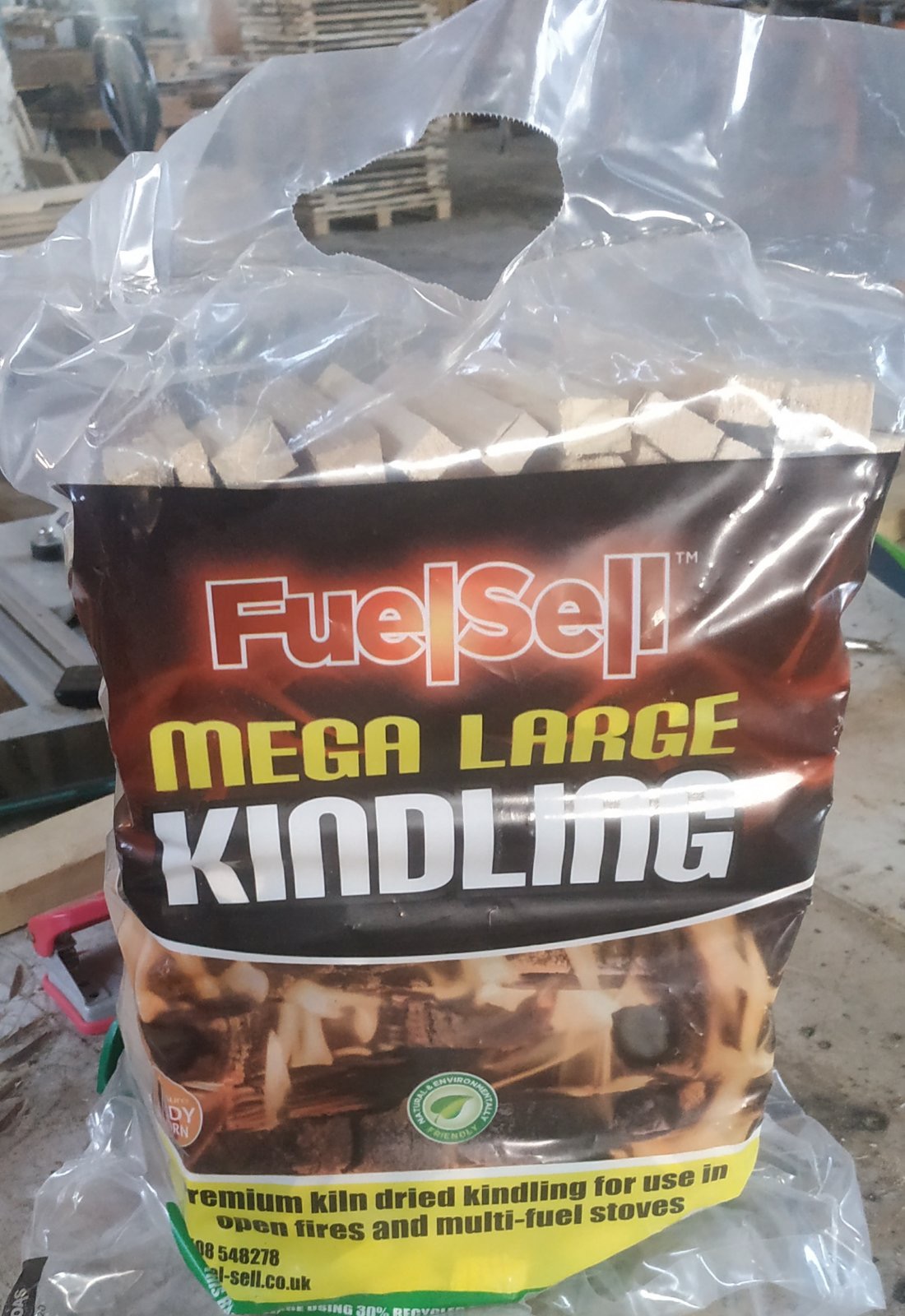 Worek z przezroczystego tworzywa wypełniony drobnymi kawałkami drewna opałowego, etykieta FuelSell Mega Large Kindling, drewniane palety w tle.