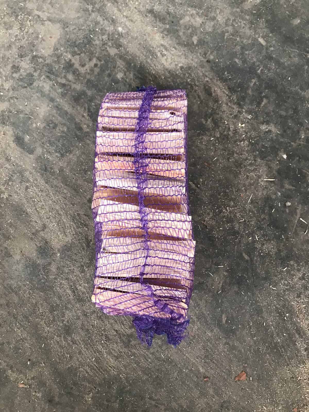 Paczka pociętego drewna kominkowego, owinięta fioletową siatką, leżąca na szarej, betonowej powierzchni.