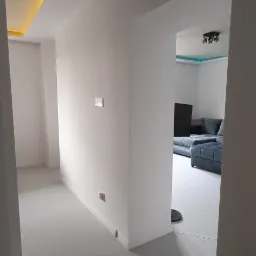 Wnętrze po odświeżeniu: pomalowane na biało ściany i podłoga w odcieniach szarości, widoczny fragment salonu z szarą sofą w tle, sufit z podświetleniem LED.