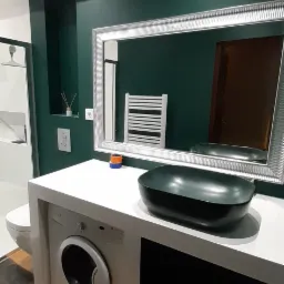 Nowoczesna łazienka z umywalką nablatową w kolorze ciemnej zieleni, białą szafką z wbudowaną pralką, lustrem w srebrnej ramie i ścianami pomalowanymi na ciemny odcień zieleni.