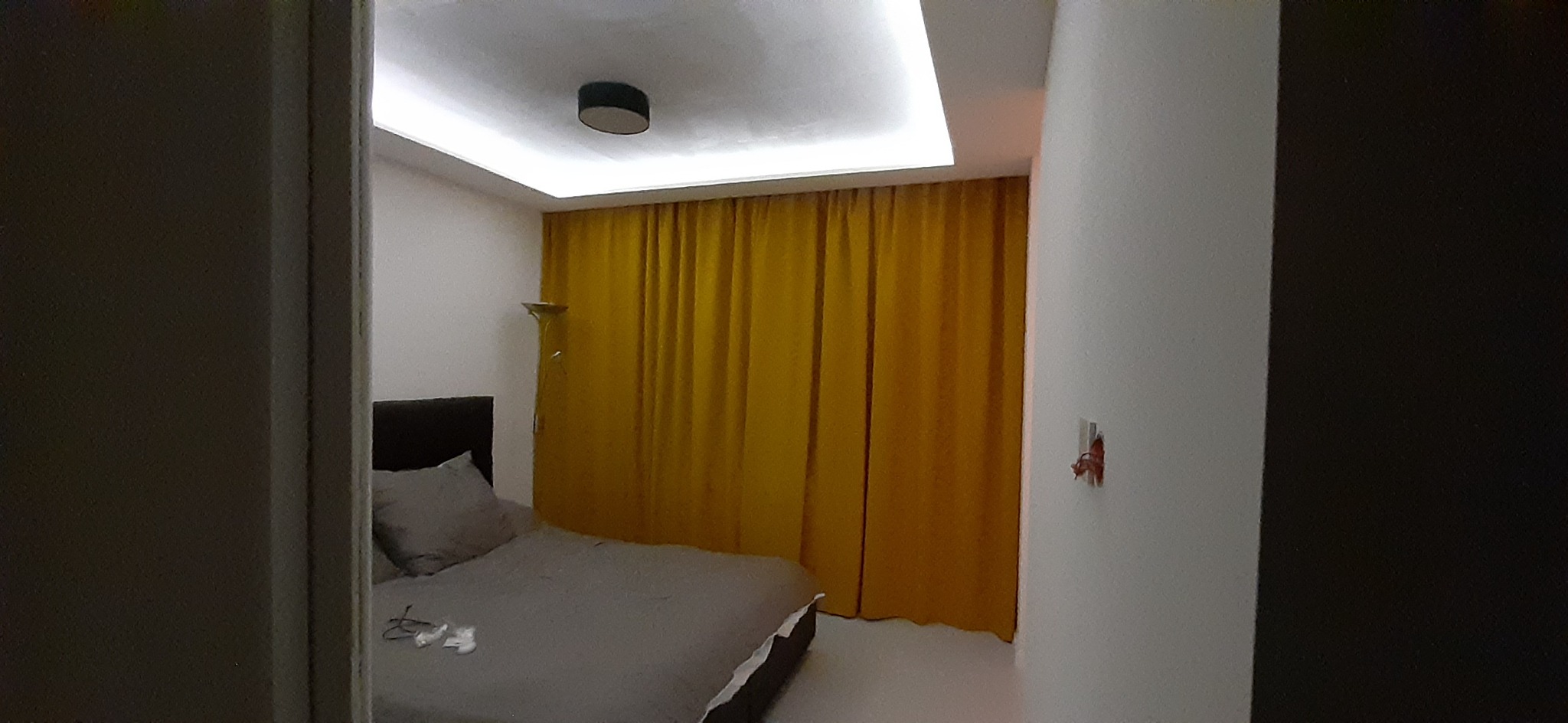 Pokój po remoncie: świeżo pomalowane ściany w odcieniach bieli, żółte zasłony, nowoczesne oświetlenie sufitowe i minimalistyczny wystrój sypialni z szarym łóżkiem.