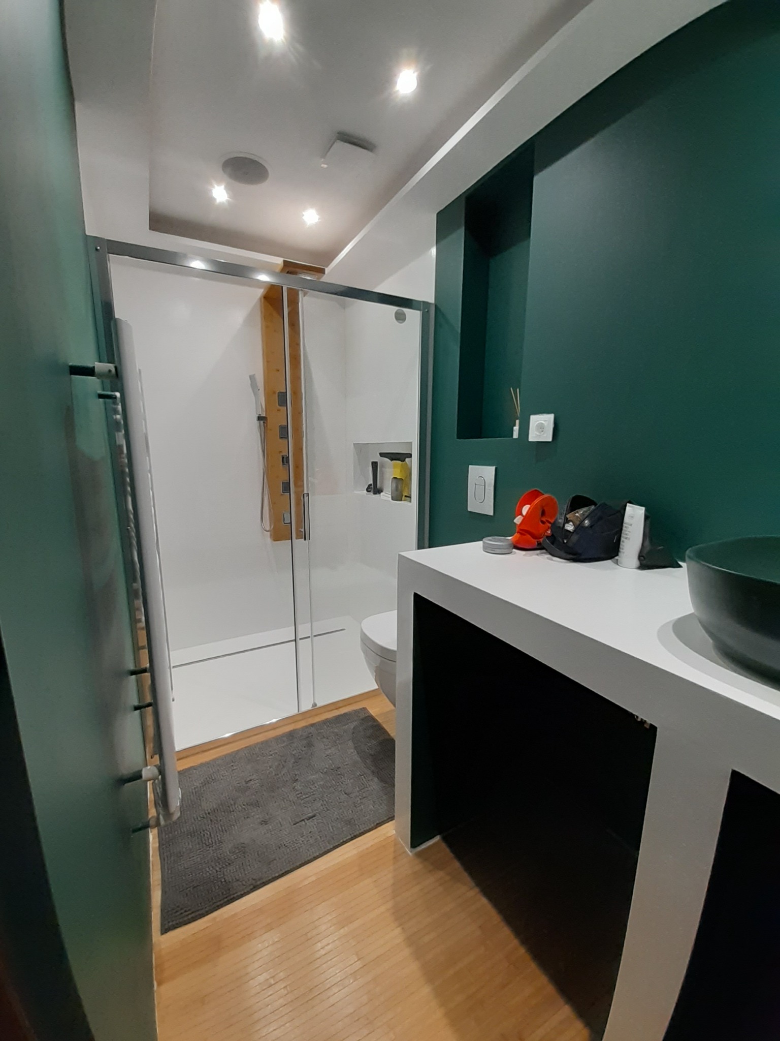 Nowoczesna łazienka z zielonymi ścianami, szklaną kabiną prysznicową, drewnianą podłogą i białą umywalką z czarną misą. Na blacie umywalki leżą kosmetyki i akcesoria.