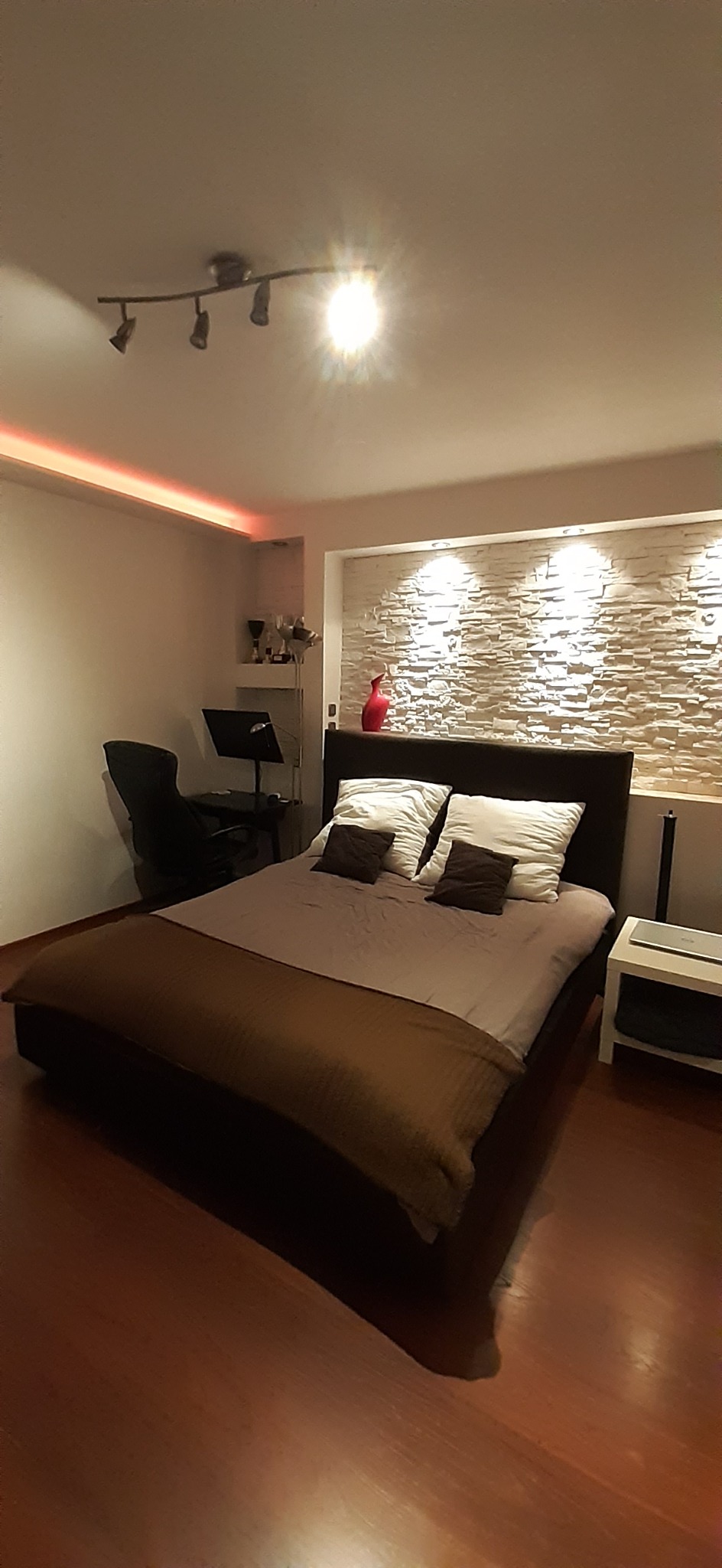 Nowoczesna sypialnia z podświetleniem LED nad łóżkiem, dekoracyjna ściana z kamienia, drewniana podłoga i lampa sufitowa z czterema reflektorami.