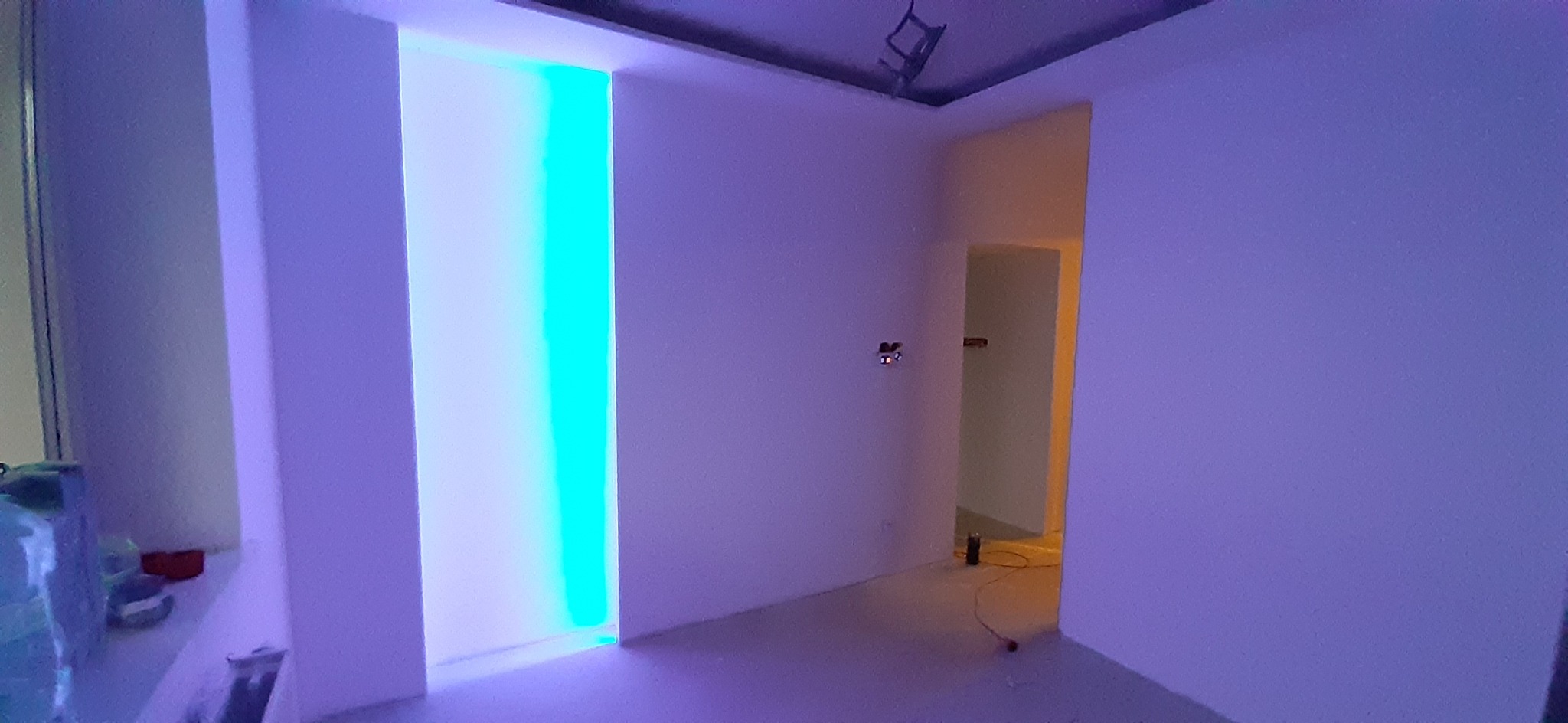 Wnętrze pomalowanego pokoju z podświetlaną wnęką w ścianie, widoczne gniazdka elektryczne i otwór drzwiowy do kolejnego pomieszczenia.