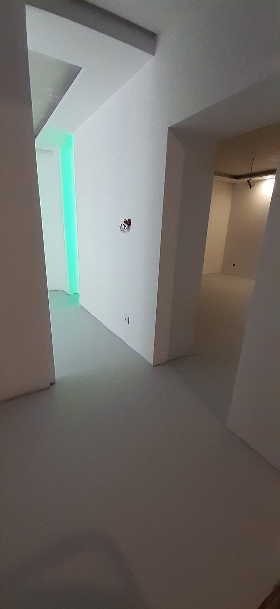 Wnętrze w stanie deweloperskim po świeżo wykonanych pracach wykończeniowych, widoczne białe ściany, przygotowane gniazdka elektryczne i oświetlenie LED w sąsiednim pomieszczeniu.