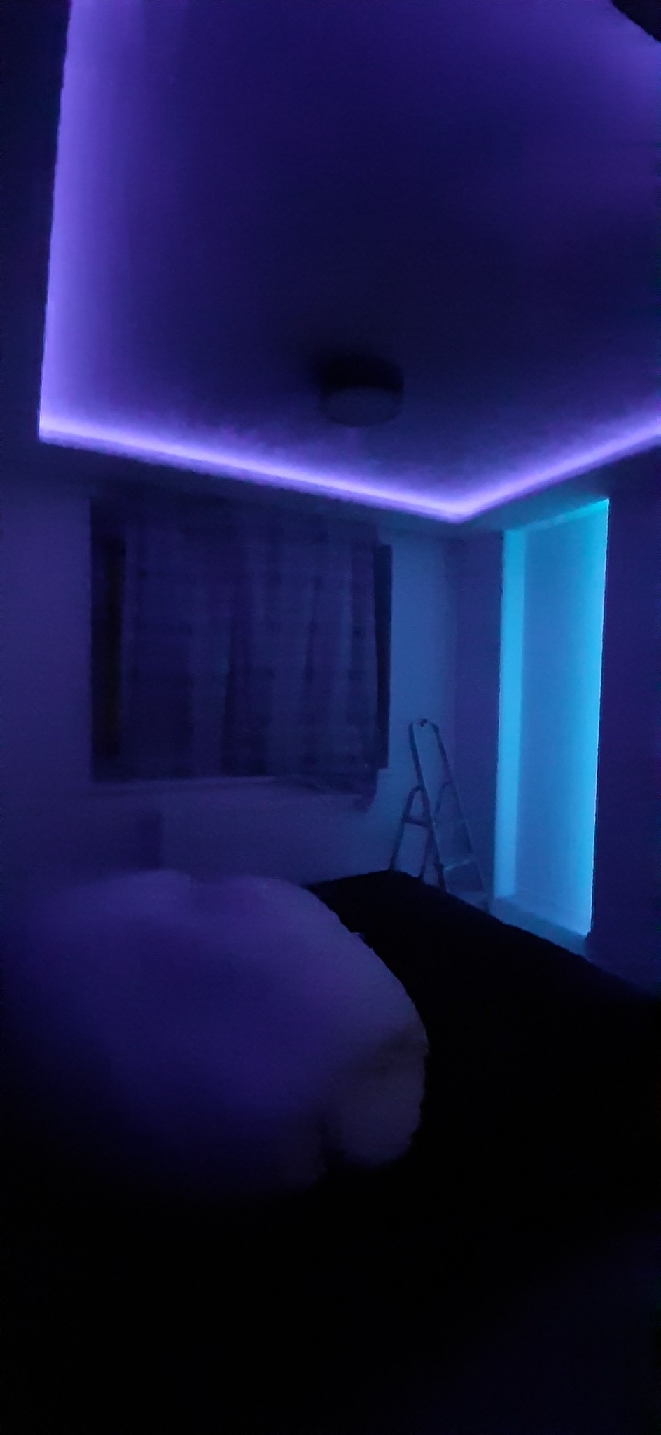 Pokój z podświetlonym sufitem i wnęką okienną w odcieniach fioletu i błękitu, drabina oparta o ścianę, widoczny fragment łóżka.