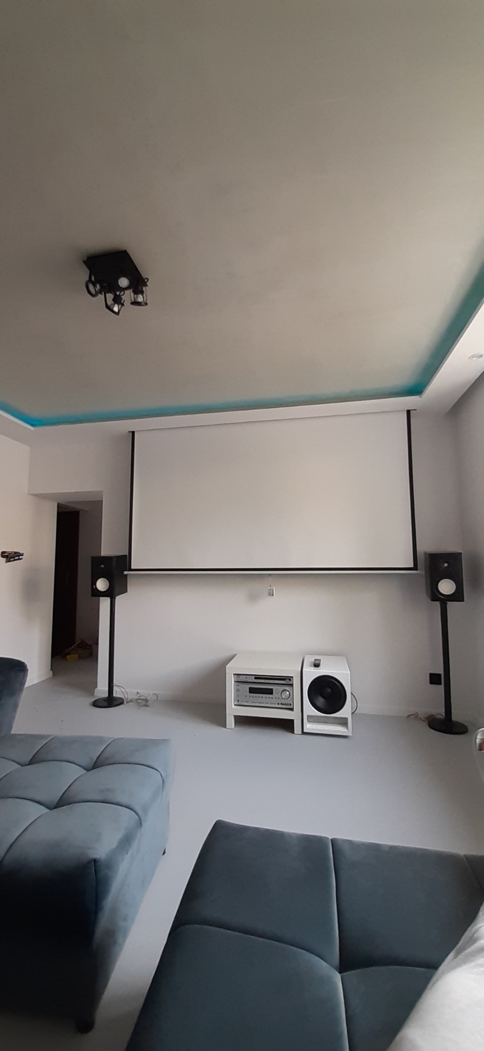 Nowoczesne wnętrze pokoju z szarymi ścianami, sufitem z oświetleniem LED, ekranem projekcyjnym, głośnikami na stojakach i niebieską, pikowaną sofą.