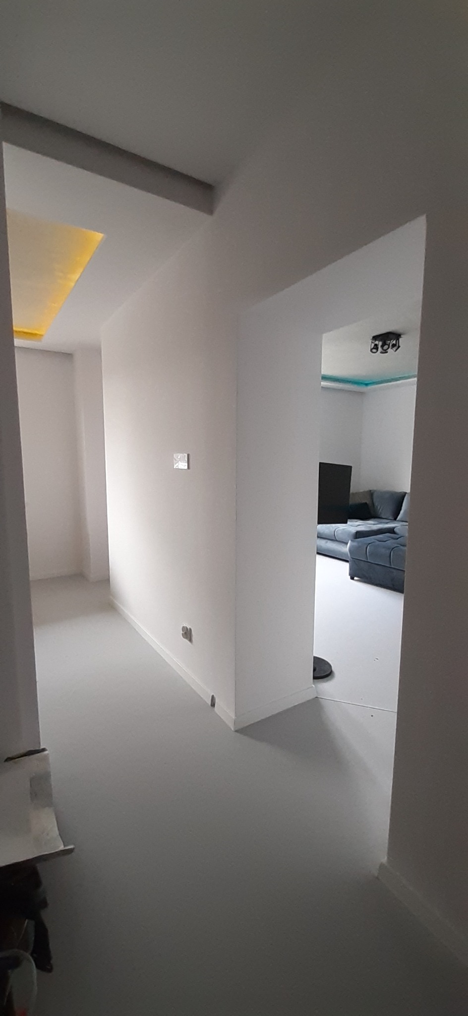 Wnętrze po odświeżeniu: pomalowane na biało ściany i podłoga w odcieniach szarości, widoczny fragment salonu z szarą sofą w tle, sufit z podświetleniem LED.