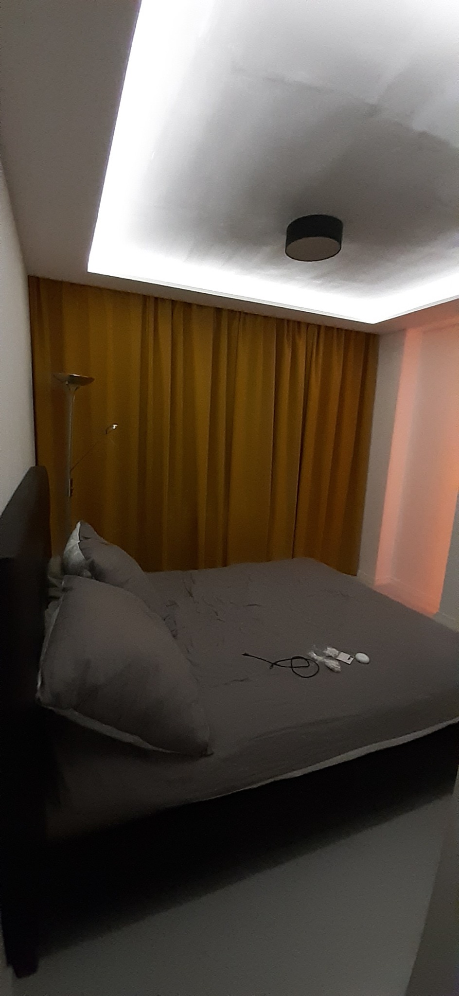 Sypialnia z szarym łóżkiem, zasłonami w kolorze musztardowym i oświetleniem LED na suficie, widoczne niedoskonałości na ścianie i suficie.