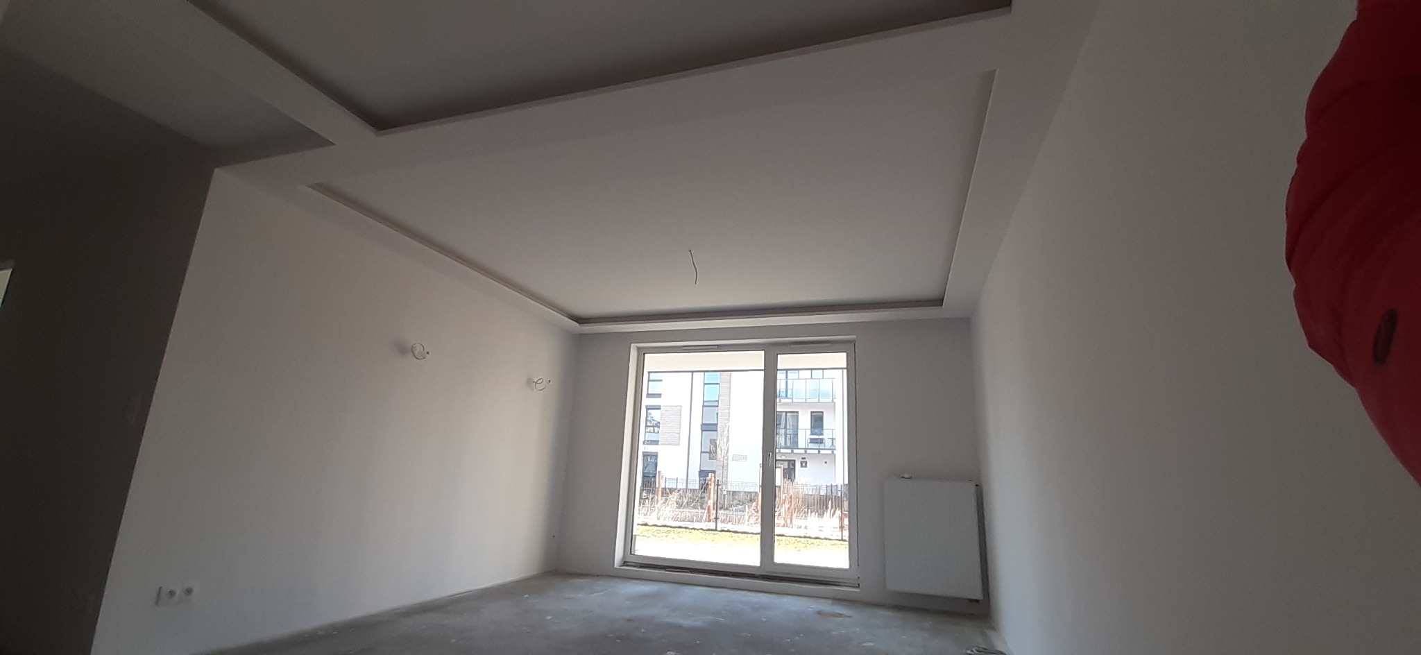 Wnętrze pokoju po remoncie z białymi ścianami, ozdobnym sufitem podwieszanym i dużym oknem balkonowym z widokiem na budynek mieszkalny.