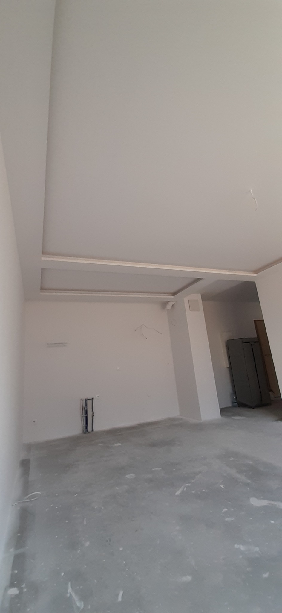 Wnętrze w stanie deweloperskim z surową podłogą i świeżo pomalowanymi ścianami na biało, widoczna instalacja hydrauliczna w ścianie oraz ozdobny sufit podwieszany z listwami.