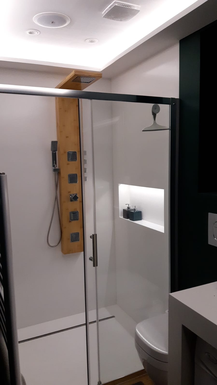 Nowoczesna kabina prysznicowa z drewnianym panelem natryskowym, szklanymi drzwiami przesuwnymi i wnęką z podświetleniem na kosmetyki w wykończonej łazience.