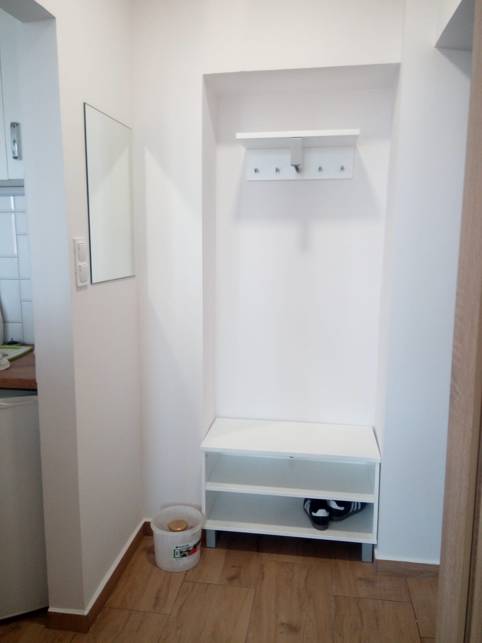 Białe, nowocześnie urządzone wnętrze przedpokoju z zabudowaną szafą wnękową, lustrem i drewnianą podłogą. Na dolnej półce widoczne buty, obok wiadro po farbie.