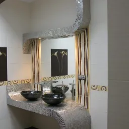 Wykończona łazienka z dwoma umywalkami wpuszczonymi w blat wykończony mozaiką, nad nimi lustro z obudową również z mozaiki, na ścianie grafika z motywem kwiatowym.