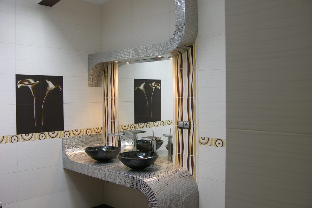 Wykończona łazienka z dwoma umywalkami wpuszczonymi w blat wykończony mozaiką, nad nimi lustro z obudową również z mozaiki, na ścianie grafika z motywem kwiatowym.