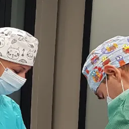 Dwoje chirurgów w kolorowych czepkach ochronnych i maskach, pochylonych nad pacjentem podczas operacji.