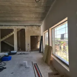 Surowe wnętrze domu w trakcie prac wykończeniowych: świeżo otynkowane ściany, widoczne kable, niebieskie rury instalacyjne i pracownik na zewnątrz budynku widziany przez okno.