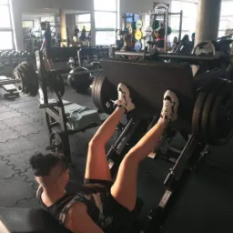 Kobieta ćwiczy na suwnicy do nóg w siłowni, widoczna z boku, w tle inne osoby i sprzęt do ćwiczeń.