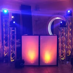 Scena z dwoma podświetlanymi panelami DJ-skimi, reflektorami na kratownicach oświetleniowych i fioletowym oświetleniem ambientowym.