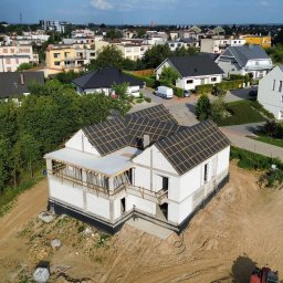 Paweł Mejza BUDOWNICTWO - Budowa domu jednorodzinnego w trakcie realizacji. Widok z drona na budynek z białego pustaka z częściowo ułożoną konstrukcją dachu na tle zabudowy miejskiej.