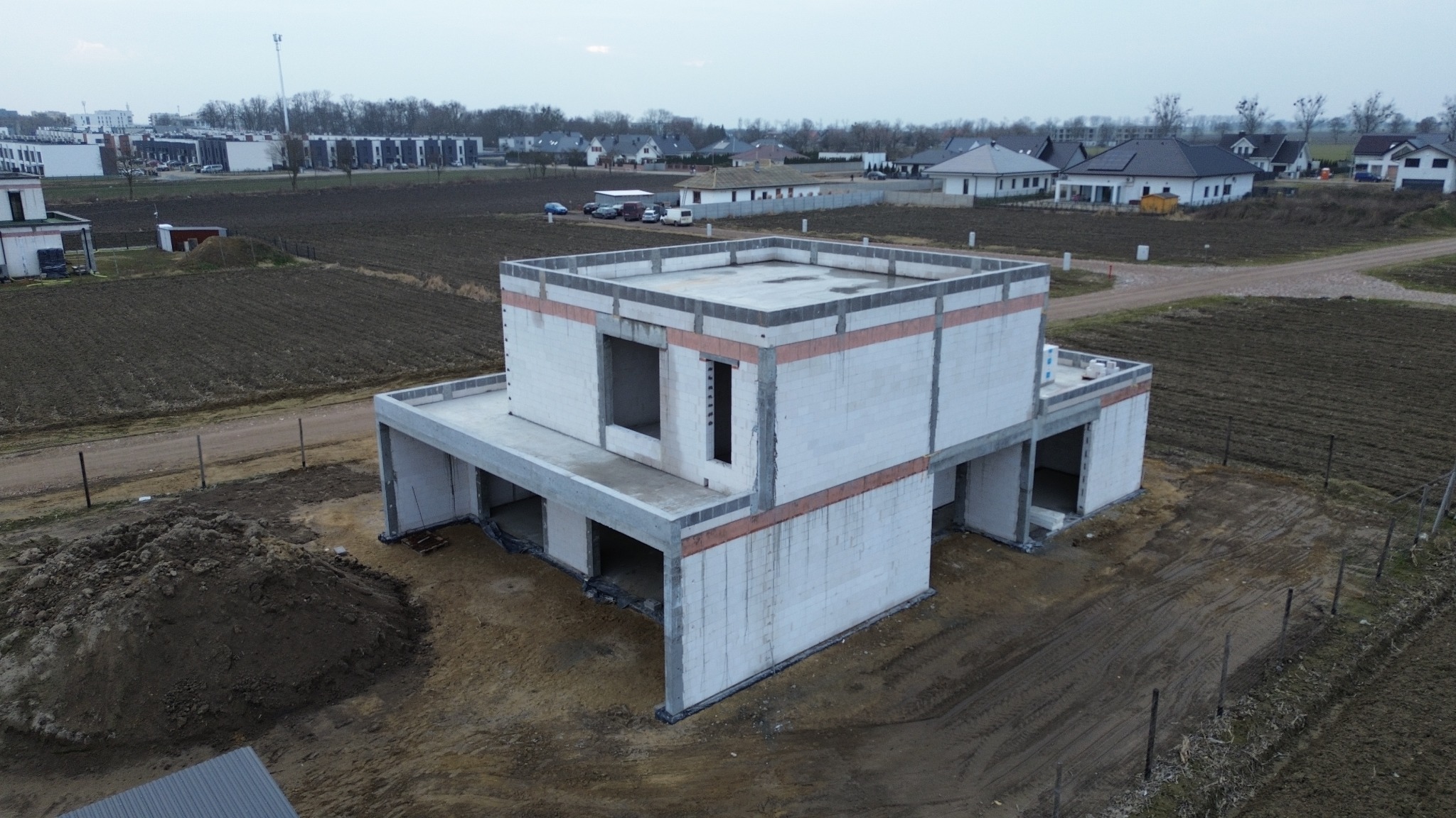 Surowy stan dwukondygnacyjnego domu z białych bloczków, widok z lotu ptaka, z widocznymi pustymi otworami okiennymi, niezadaszony, z odsłoniętymi elementami konstrukcyjnymi, na tle zaoranej ziemi...