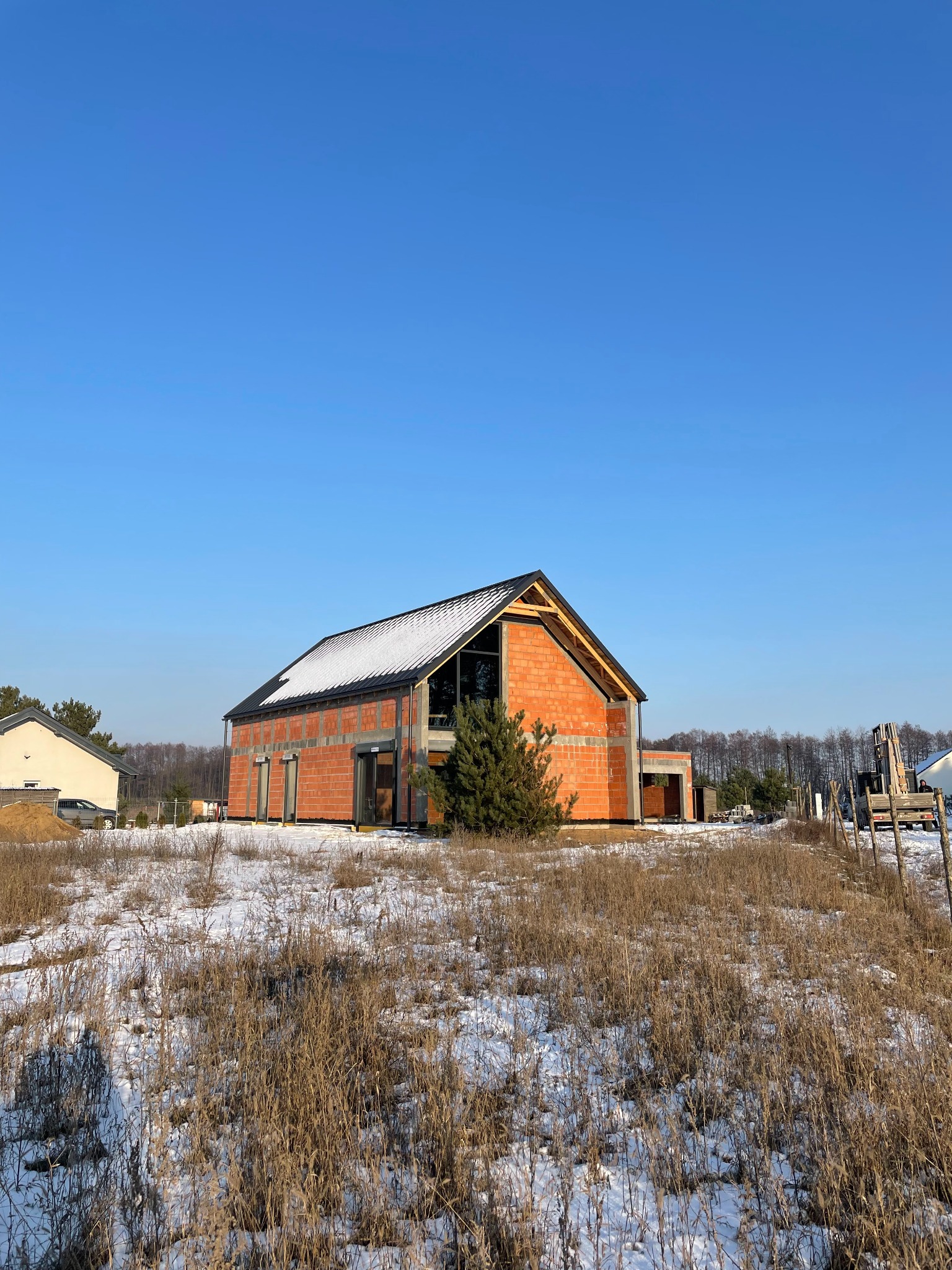 Niedokończony dom z czerwonej cegły w zimowym krajobrazie, z widocznym rusztowaniem i częściowo ośnieżonym dachem, otoczony suchą trawą i drzewami w tle.