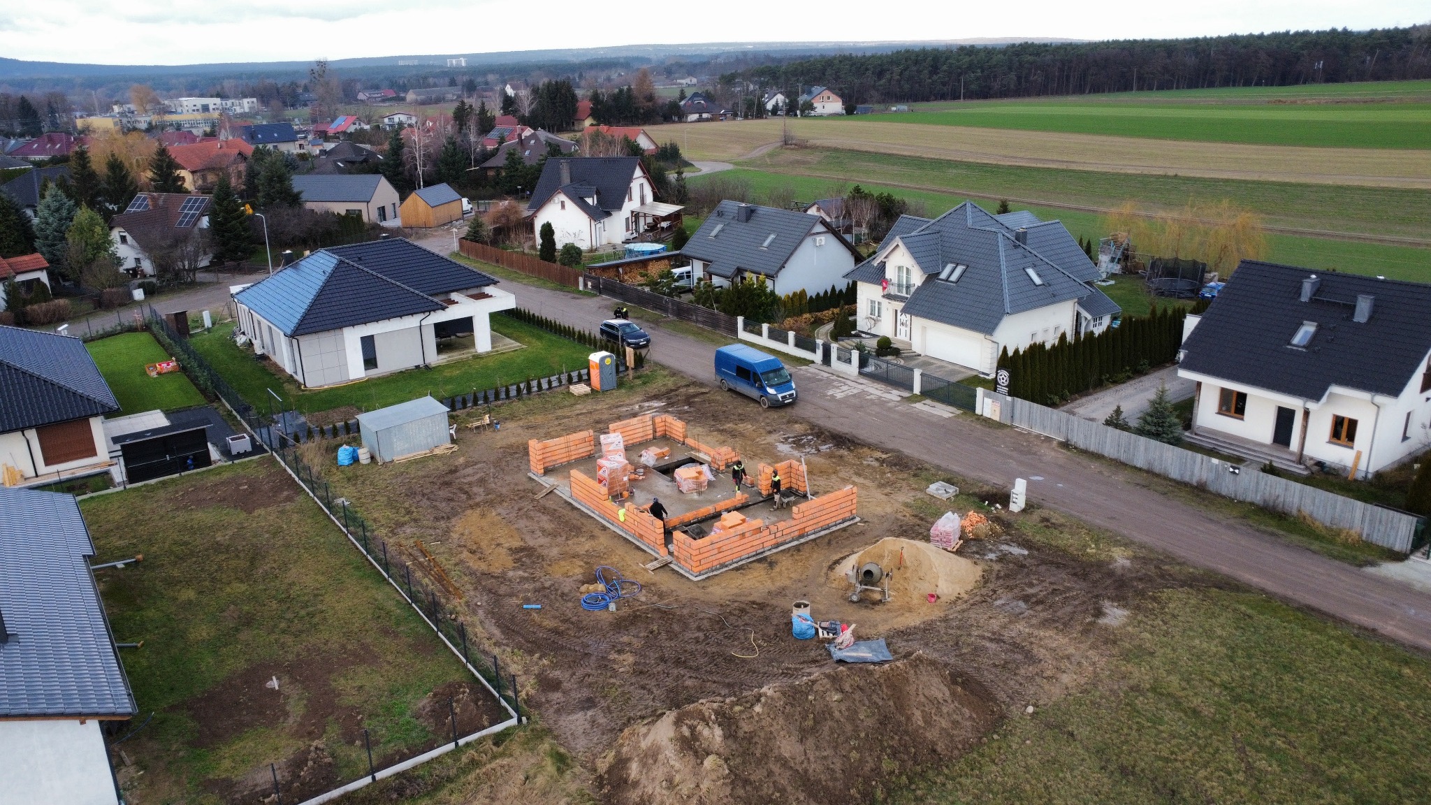 Widok z drona na plac budowy domu jednorodzinnego z czerwonej cegły, z widocznymi pracownikami, cementarką i stosem cegieł, na tle zabudowy jednorodzinnej i pól uprawnych.