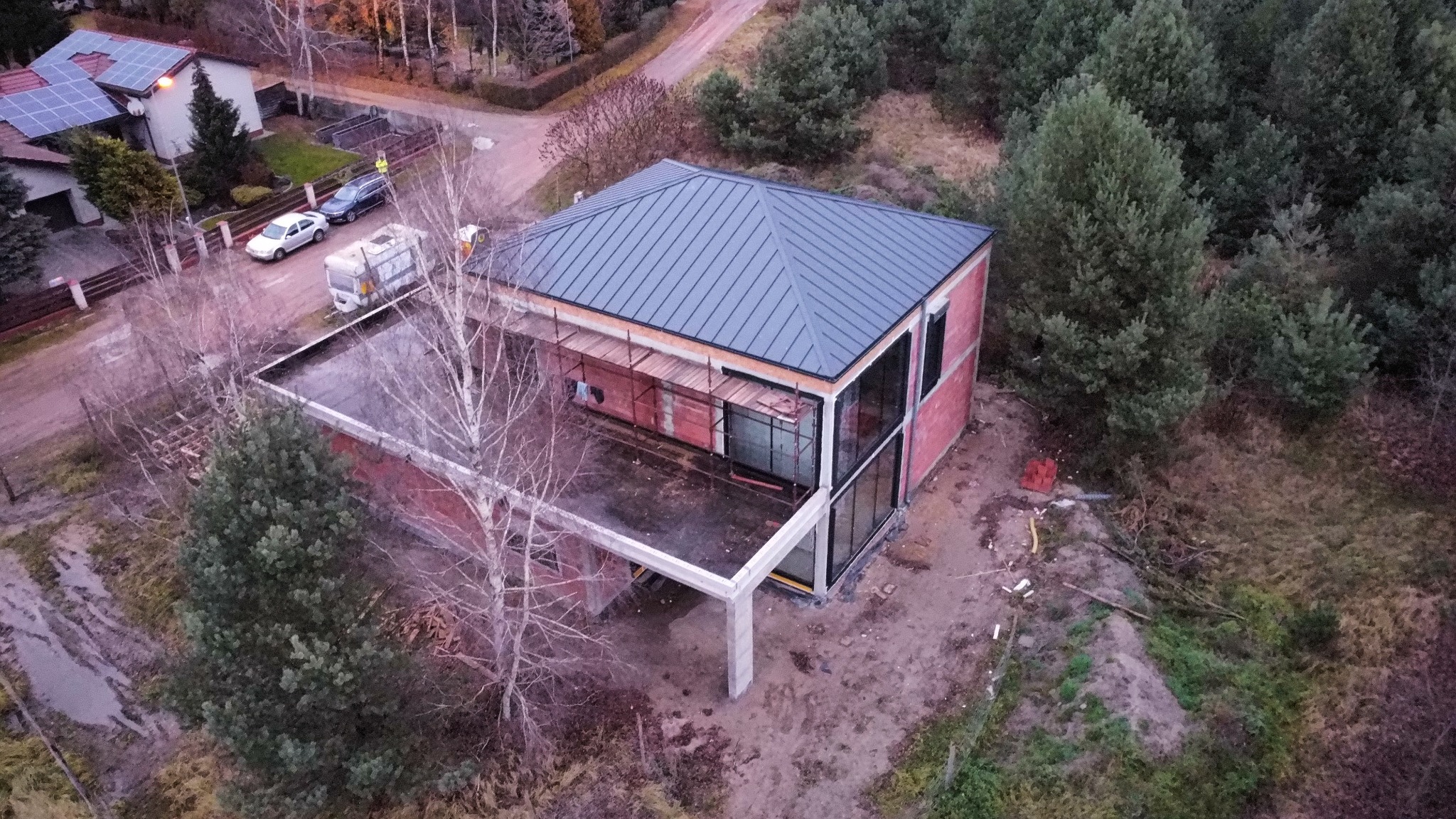 Budowa domu z płaskim dachem i dużymi oknami, widok z góry na częściowo wykończony budynek z rusztowaniami i betonowym zadaszeniem, otoczony drzewami.