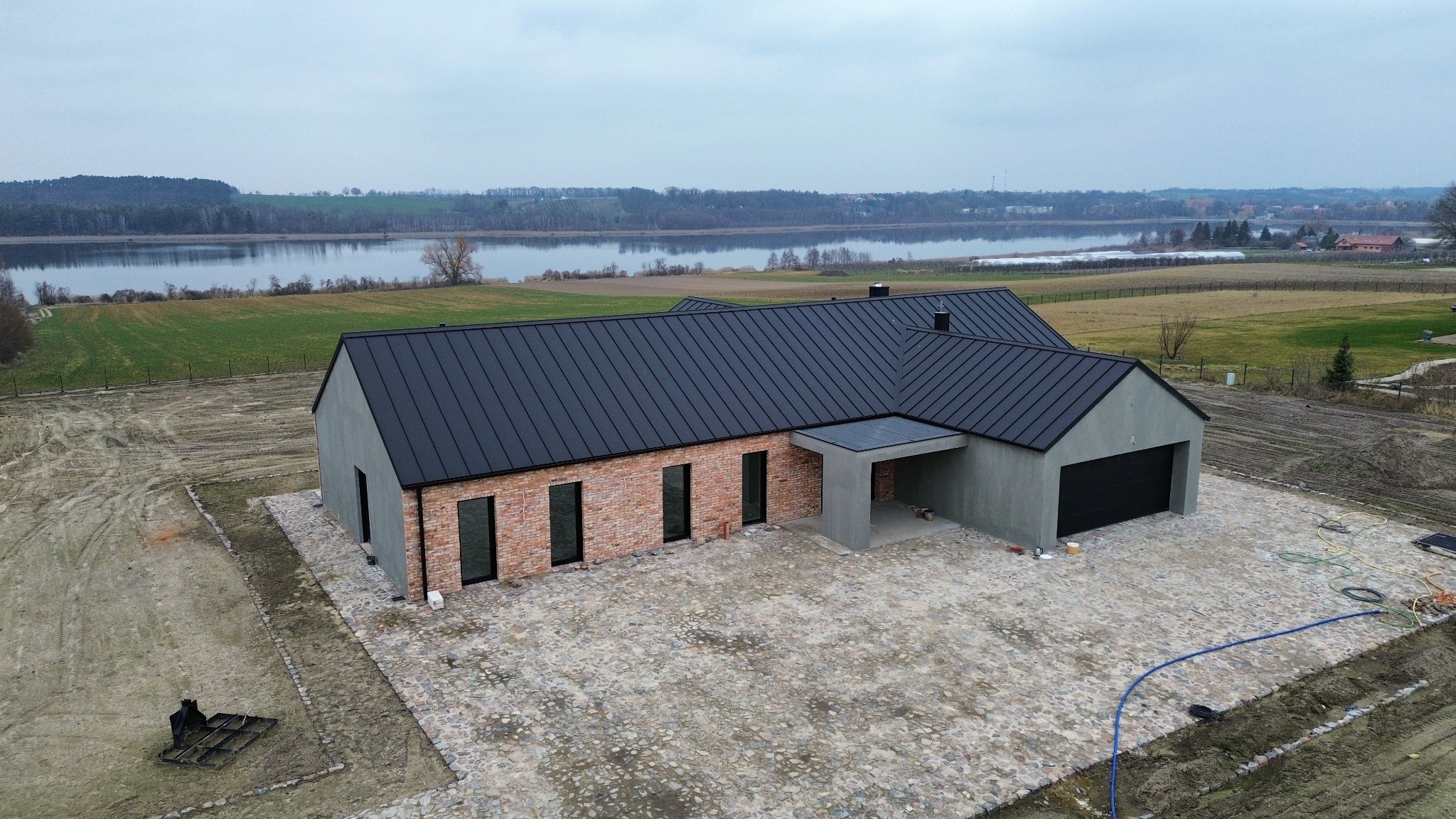 Nowoczesny dom z ciemnym dachem i ceglaną elewacją, widok z góry na tle jeziora i pól. Uwagę przykuwa kamienny podjazd i minimalistyczny design budynku.