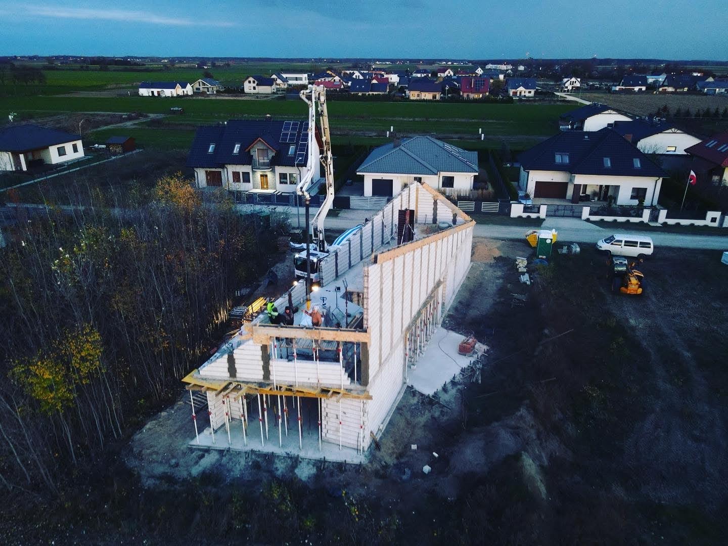Budowa domu jednorodzinnego z betonu komórkowego, widok z drona na fundamenty i ściany parteru, pompa do betonu w akcji, otoczenie: zabudowa jednorodzinna i pola.