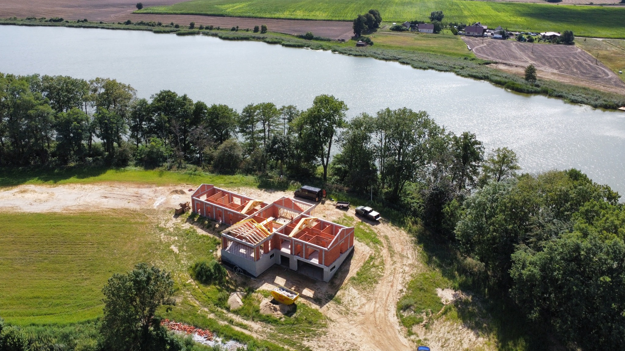 Budowa domu jednorodzinnego w Kórniku, widok z drona. Czerwone cegły, drewniana konstrukcja dachu, malownicze otoczenie jeziora i zieleni. Etap surowy otwarty.